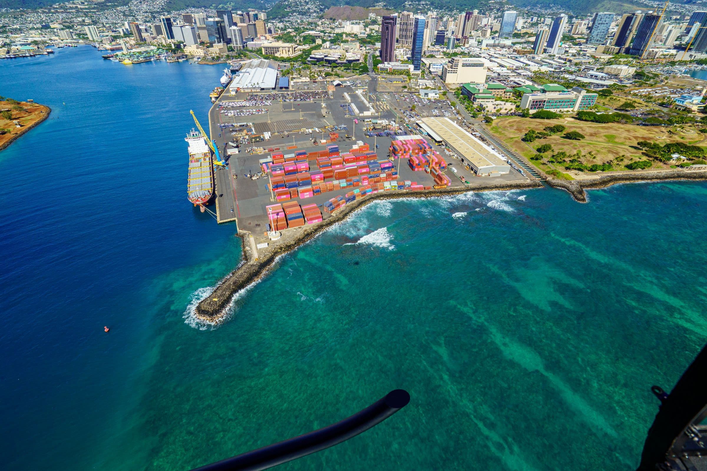 Aerial view of a cargo port on a coastline, with terminals, containers and the city beyond